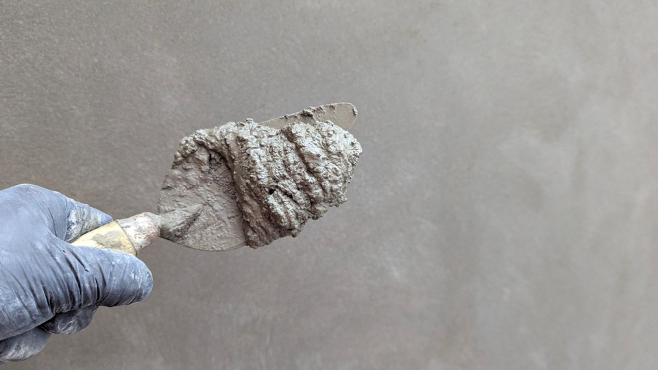 Een troffel met grijs stuc en een hand in een blauwe handschoen bij een grijze wand